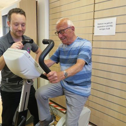 Fysiotherapeut Yordi Appelhof in zijn grijze shirt helpt meneer Wallinga in zijn blauwe shirt met sporten en bewegen met parkinson en artrose op de hometrainer