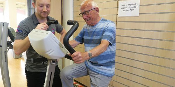 Sporten en bewegen met parkinson Fysiotherapeut Yordi Appelhof in zijn grijze shirt helpt meneer Wallinga in zijn blauwe shirt met sporten en bewegen met parkinson en artrose op de hometrainer