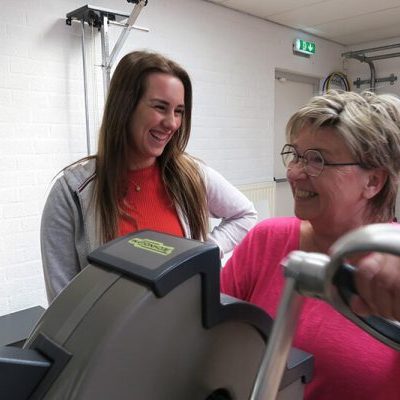 Vrouw traint bij de fysio haar armen en wordt met fysiotherapie in Meerssen geholpen door een fysiotherapeut in een grijs vest, de behandeling die ze heeft valt in de basisverzekering