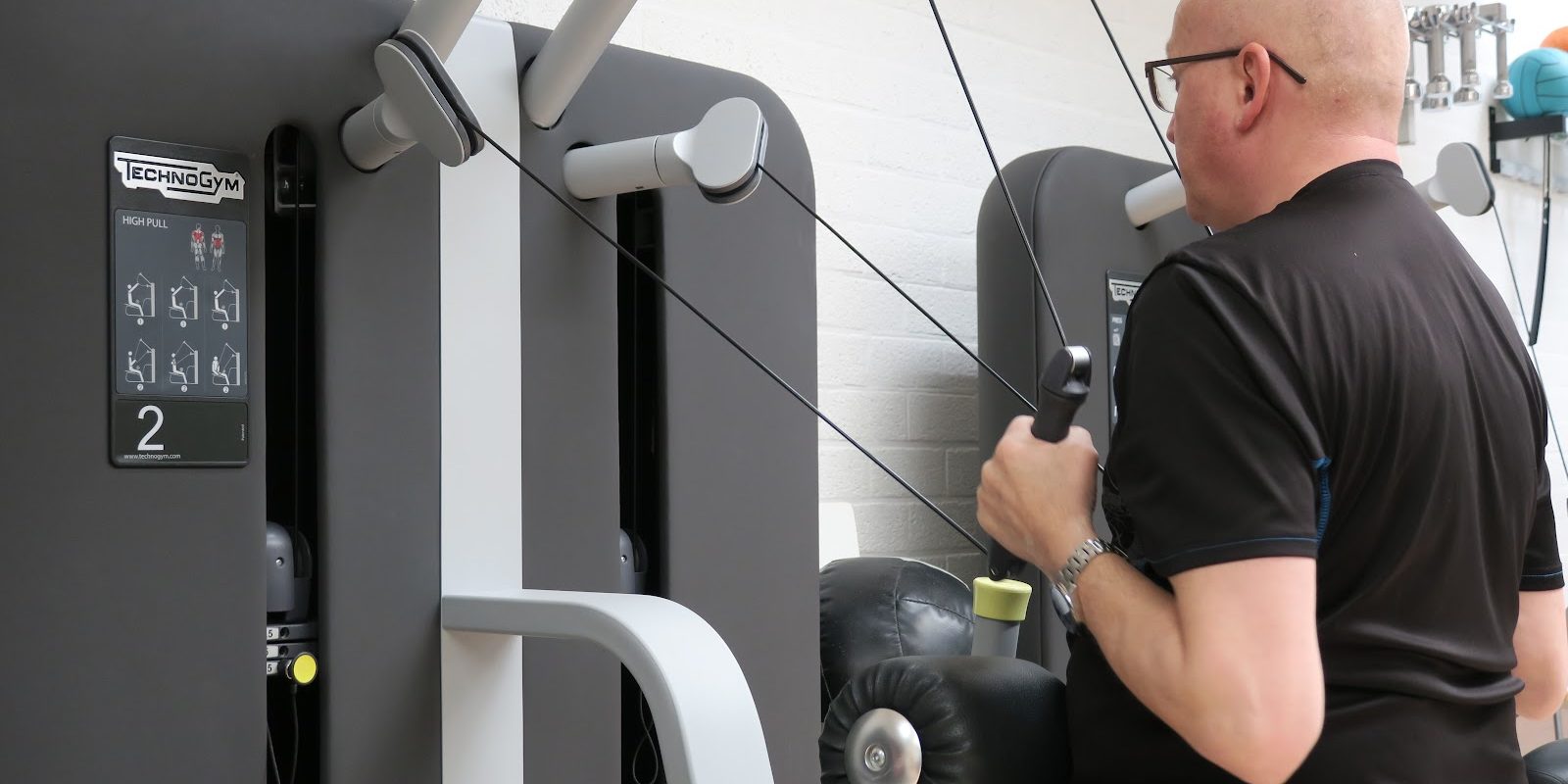 Bewegen Een man met een zwart t-shirt en een bril is aan het bewegen met behulp van een fitnessapparaat en krijgt eenfysiotherapie behandeling in Gronsveld.