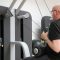 Een man met een zwart t-shirt en een bril is aan het bewegen met behulp van een fitnessapparaat en krijgt eenfysiotherapie behandeling in Gronsveld.